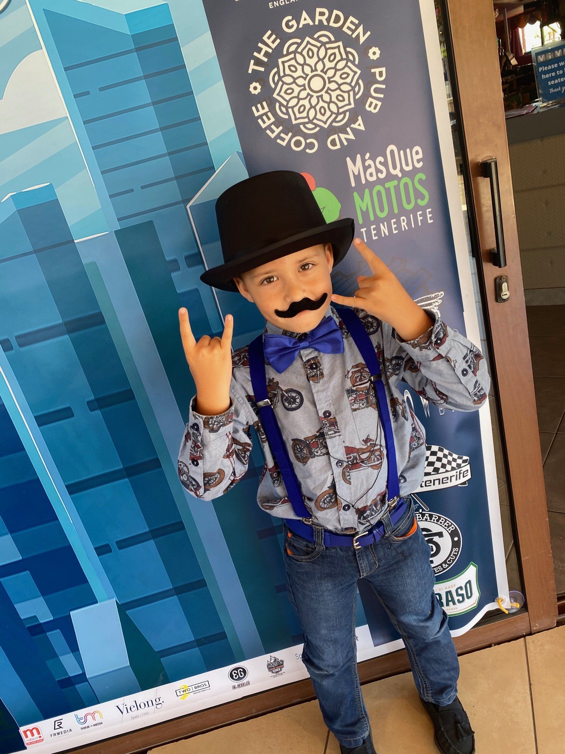 A young boy wearing a hat and suspenders is standing in front of a sign that says mas que motos