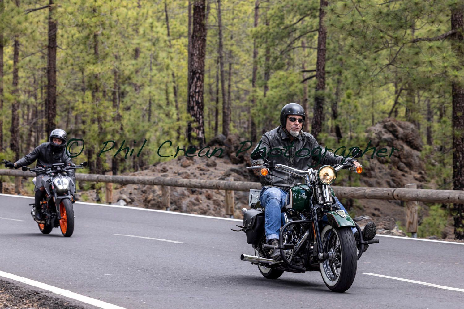 Two men are riding motorcycles down a road in the woods.