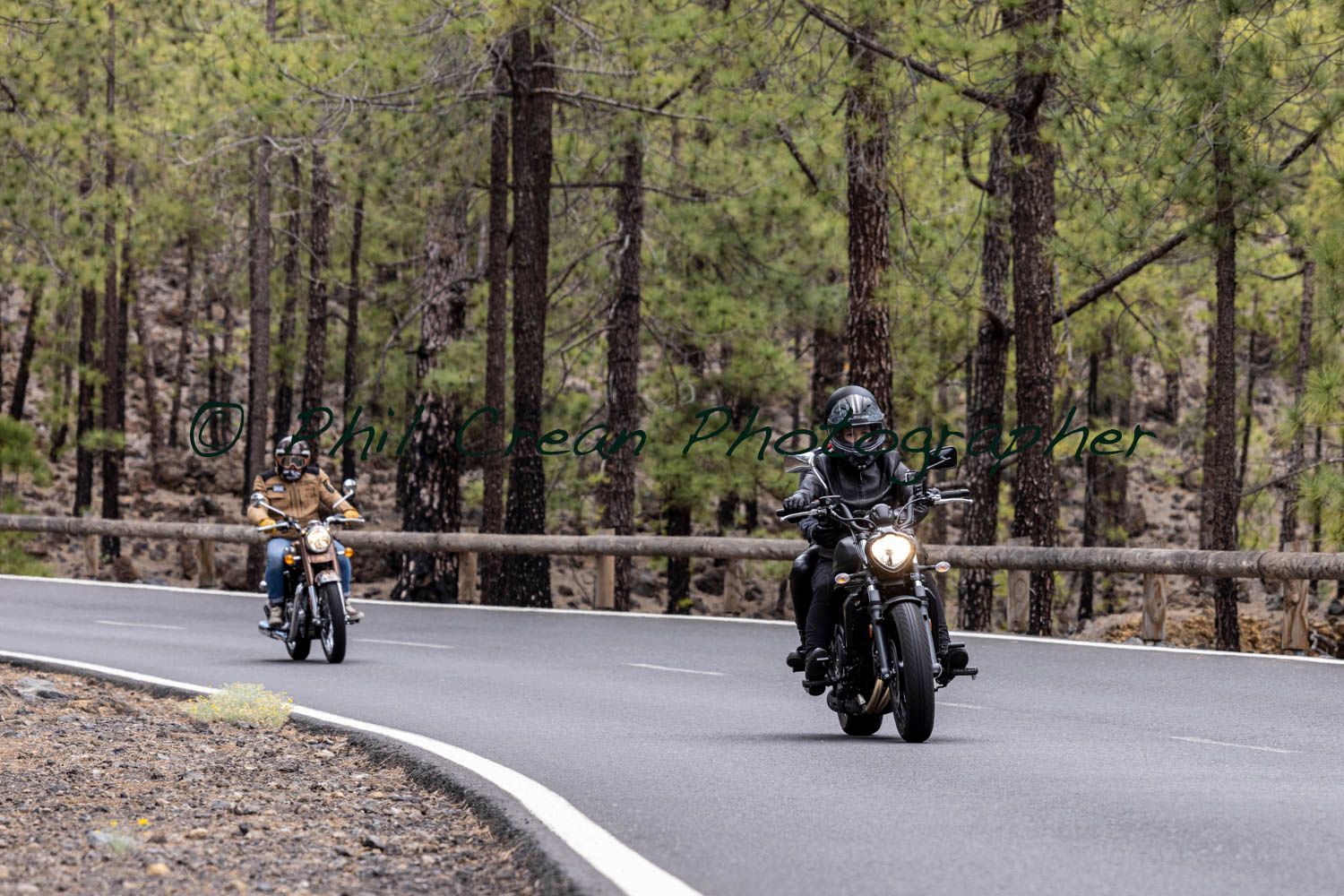 Two people are riding motorcycles down a road in the woods.