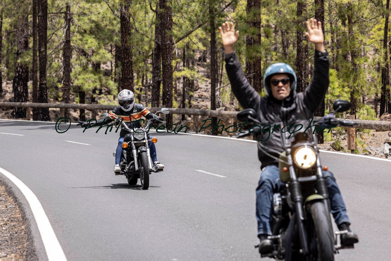 Two men are riding motorcycles down a road with their arms in the air