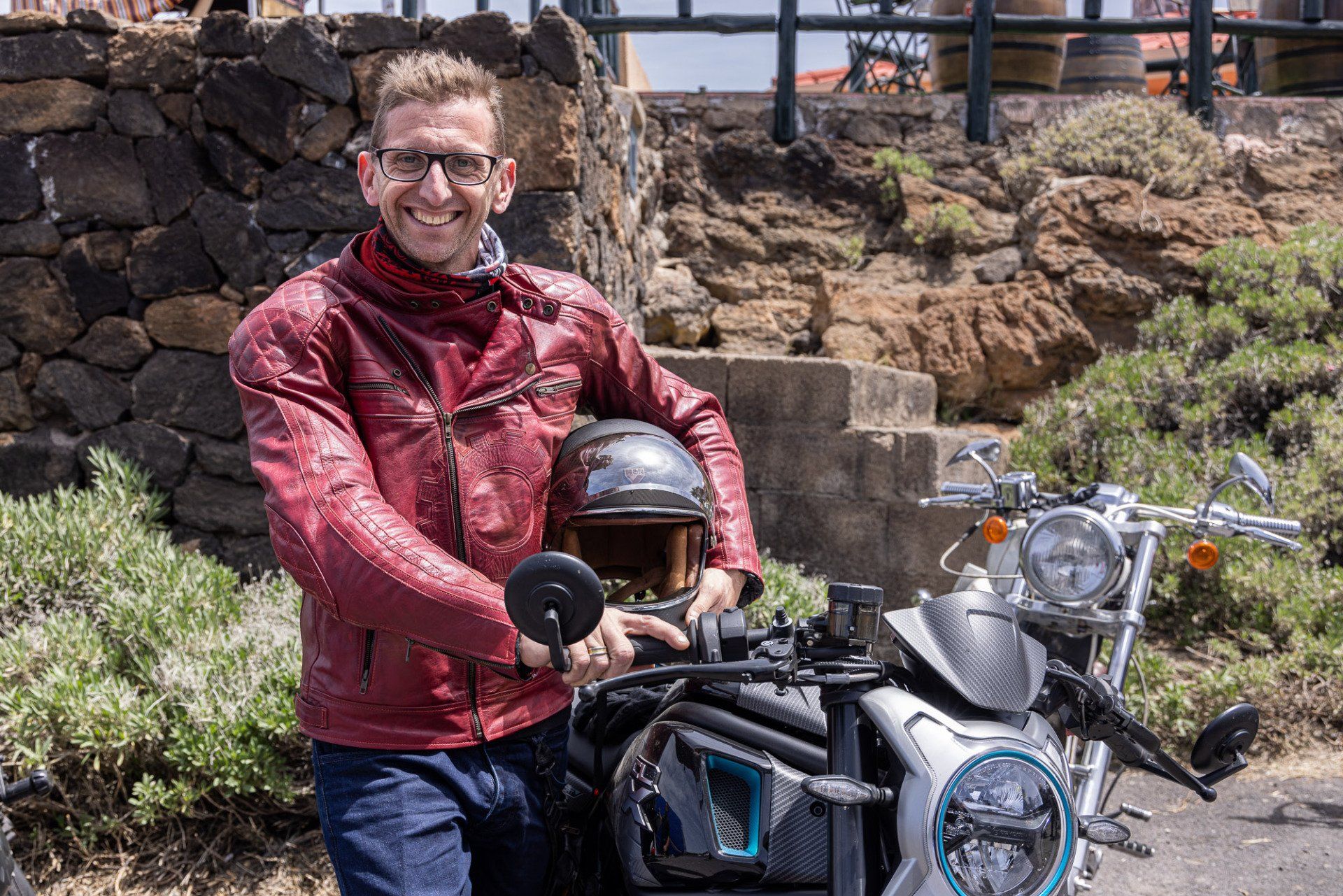 A man in a red jacket is standing next to a motorcycle.