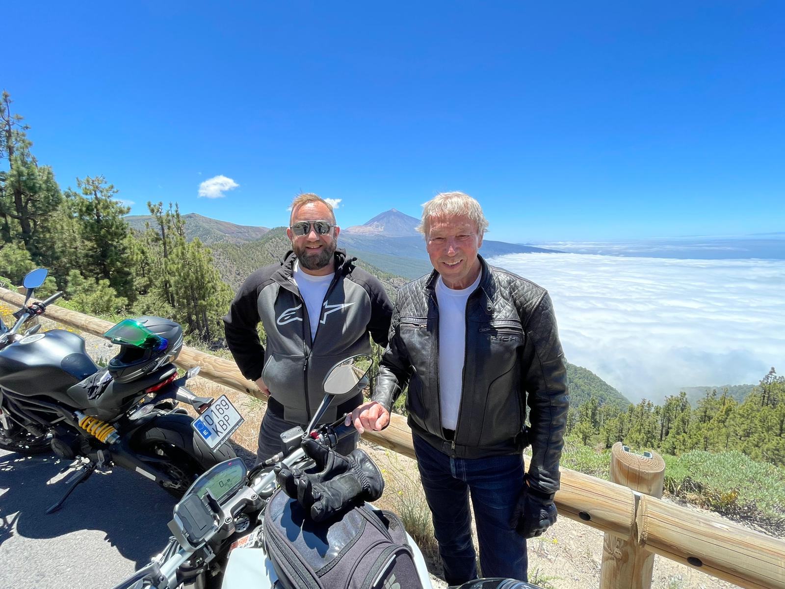 Two men are standing next to a motorcycle on top of a hill.