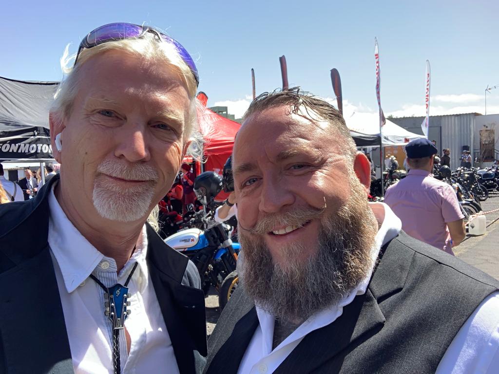 Two men with beards are posing for a picture at a motorcycle show.