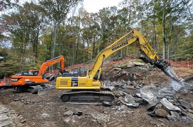 Two excavators, one orange and one yellow, break rocks in a wooded area. The yellow excavator has a demolition tool attached.