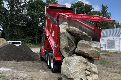 Red dump truck unloading large rocks onto a dirt lot near a pile of gravel and a partially constructed building.