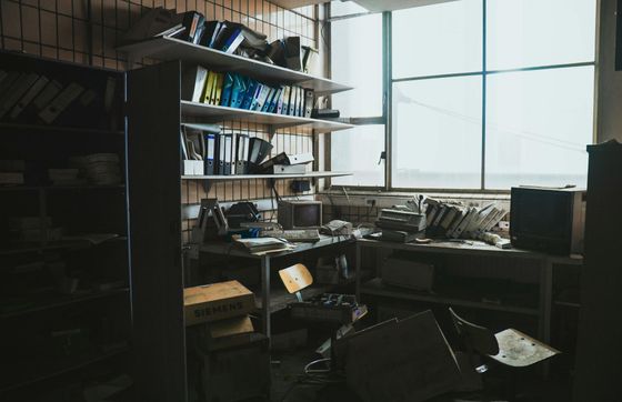 A messy office with a desk , chair , shelves and a window.