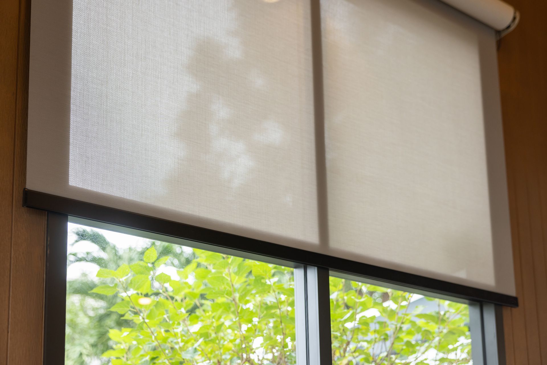 Indoor roller blinds partially lowered on a large window with greenery visible outside.
