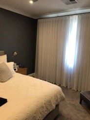 A bedroom with white sheer curtains on a large window, a grey accent wall, and a bed