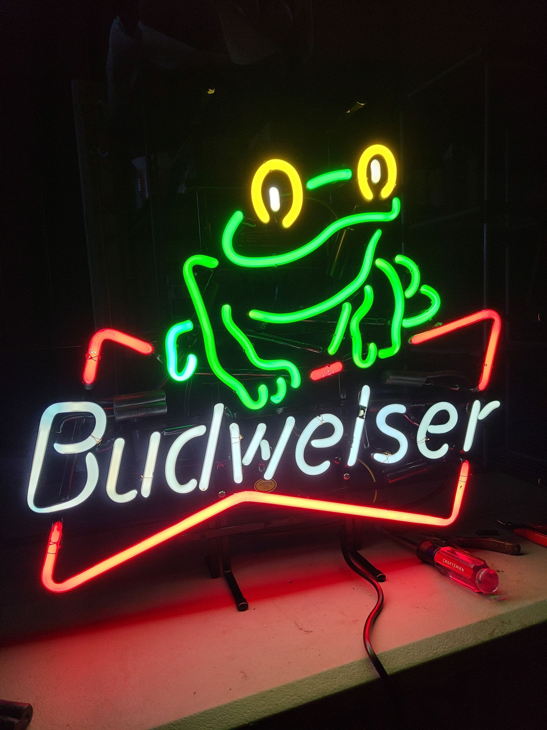 Neon Budweiser sign with green frog and white text on a red and white background.