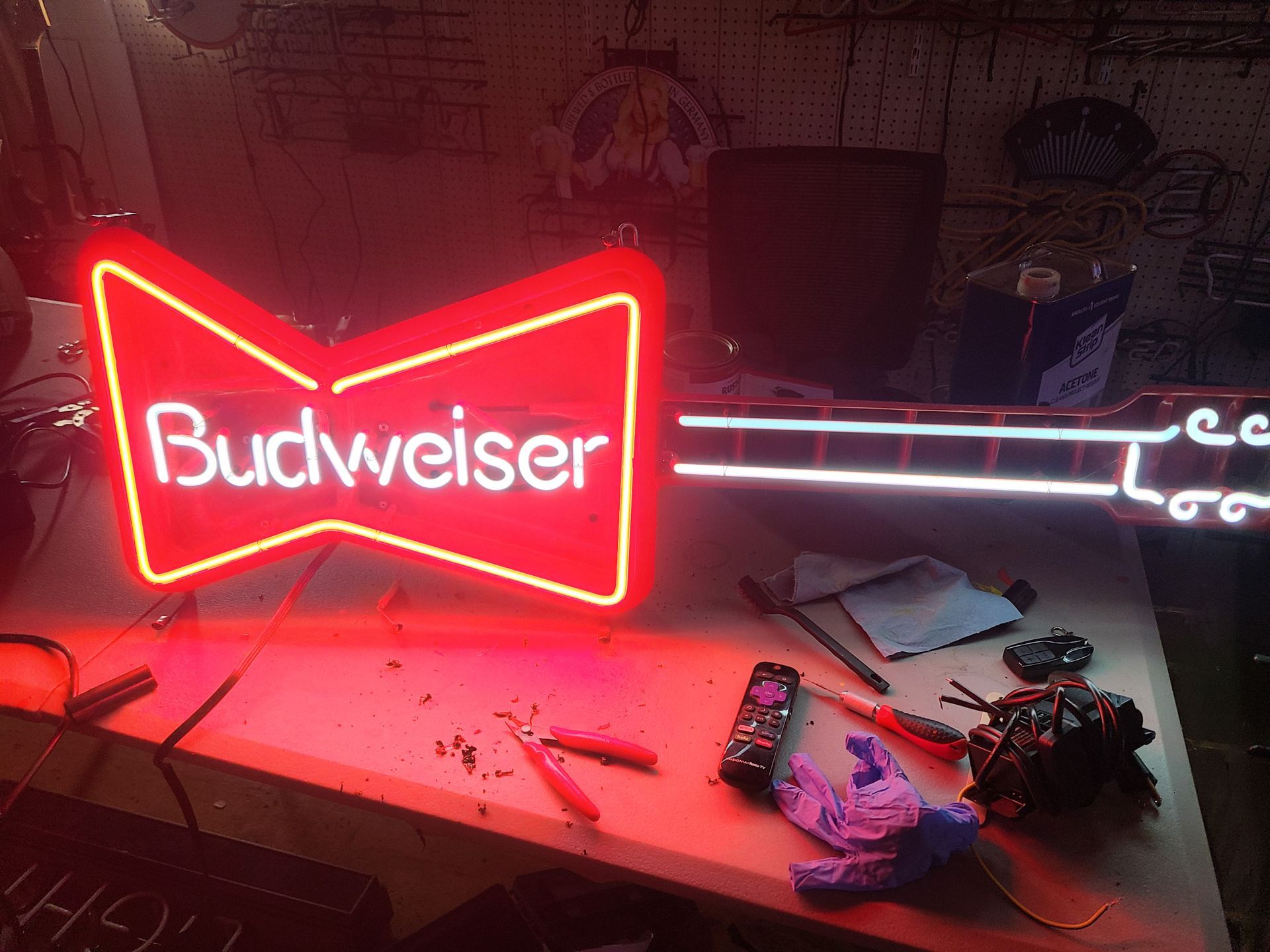 A neon Budweiser sign glows red. Tools and parts are scattered on a workbench in a dimly lit setting.