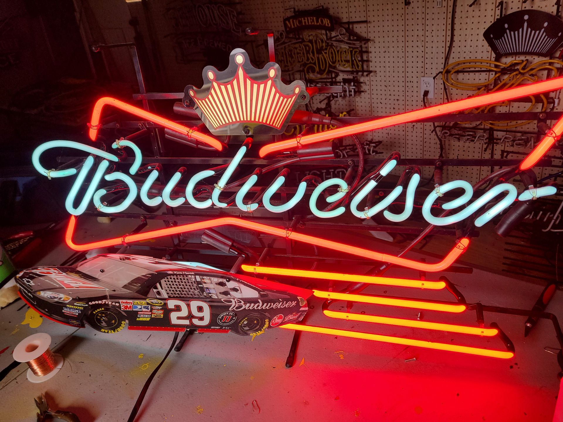 Budweiser neon sign with red and blue lettering, a crown, and a race car in a workshop setting.