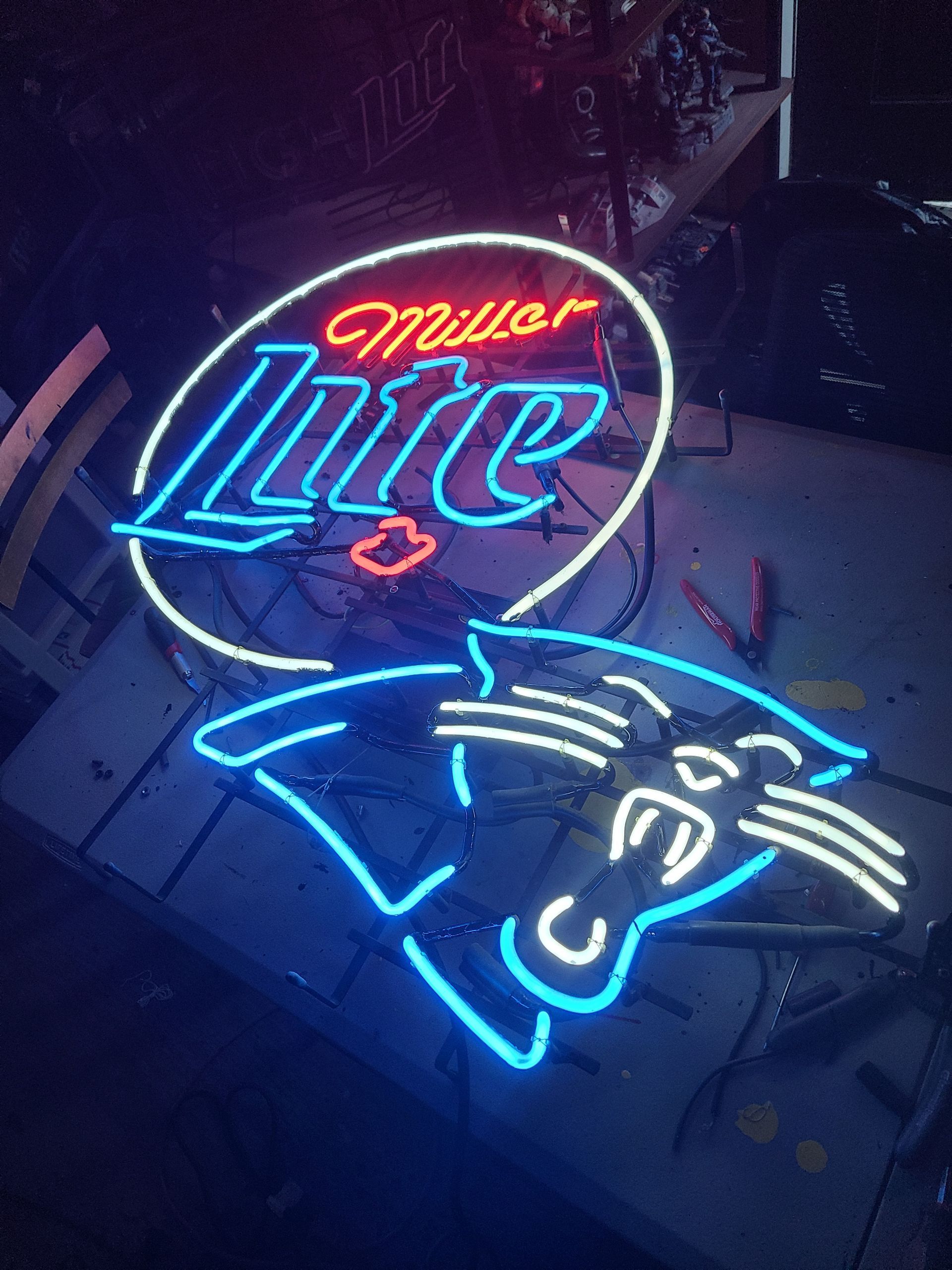 Neon Miller Lite sign above a Carolina Panthers logo, both glowing blue, white, and red in a dark setting.