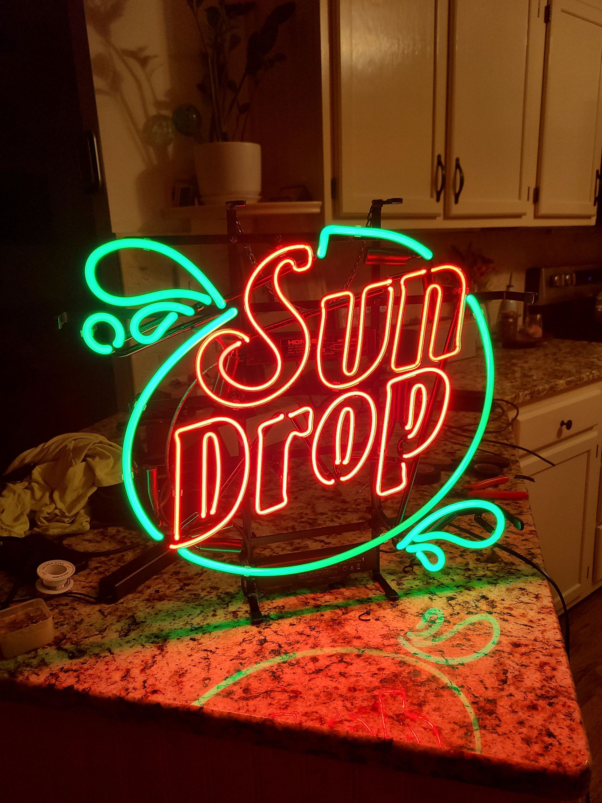 Neon Sun Drop sign, red letters within a green outline, sitting on a speckled countertop in a kitchen.