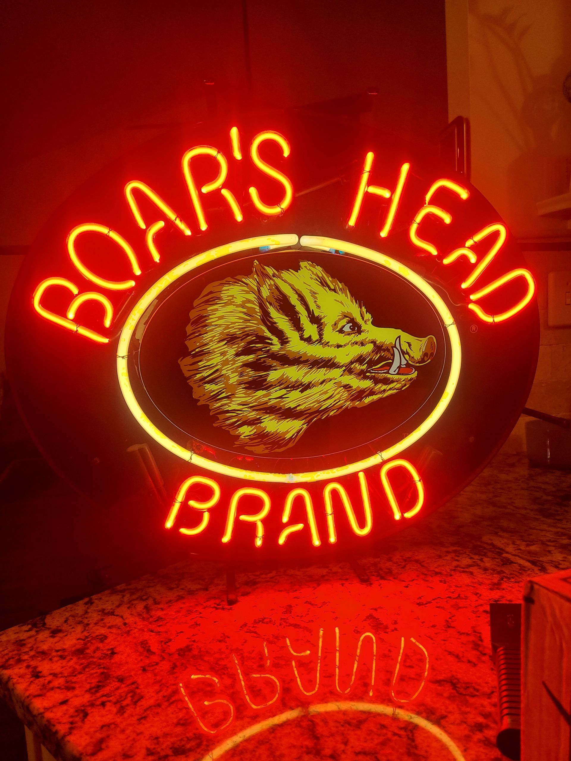 Neon Boar's Head Brand sign. Red neon lettering, yellow logo of a boar's head. Displayed on a red-lit surface.
