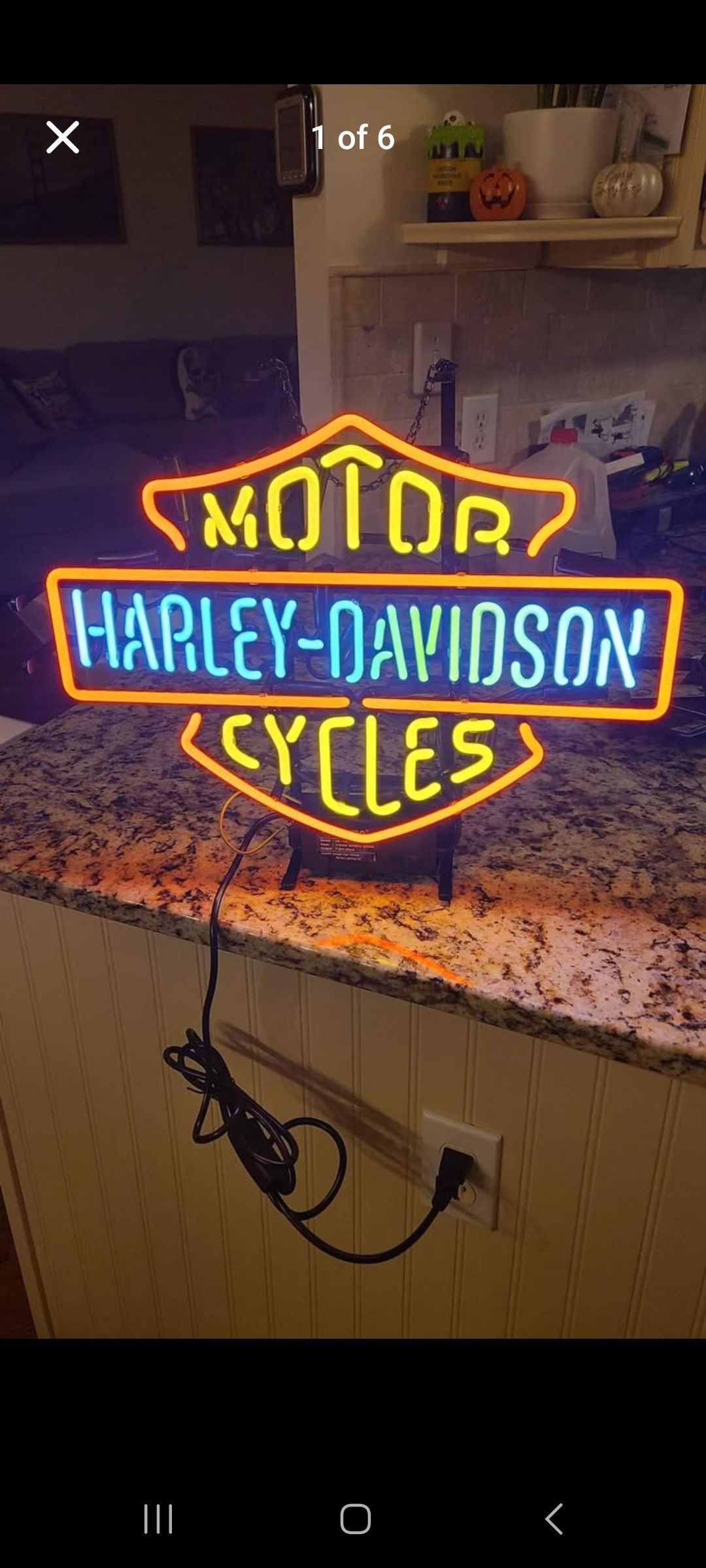 Neon Harley-Davidson sign lit up on a countertop. Black cord plugged into a wall outlet.