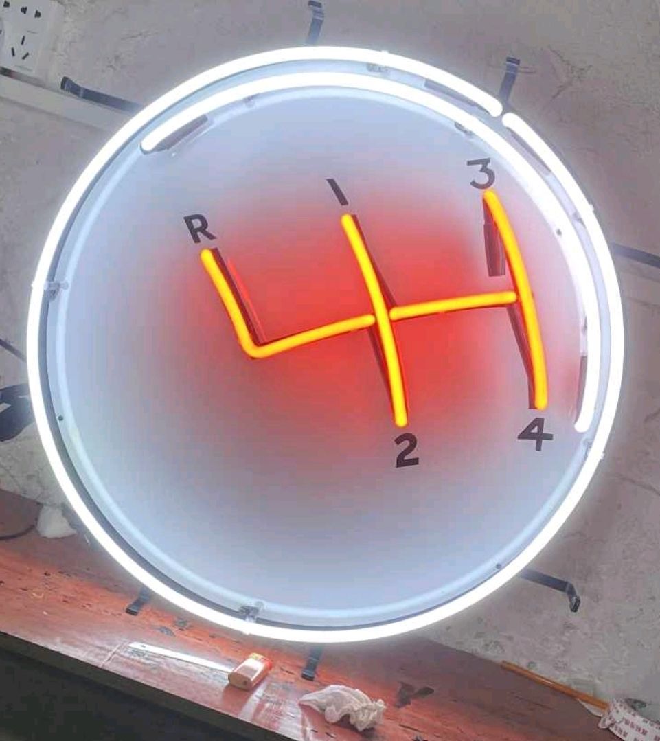 Neon sign with illuminated gear shift pattern: red and yellow on a white circle.