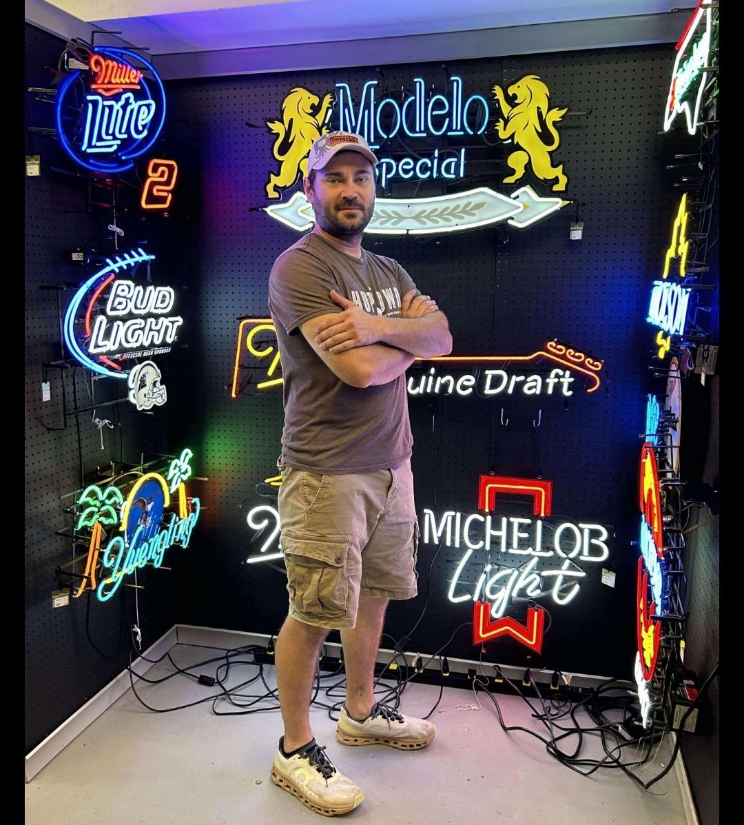 Man standing in a room with illuminated neon signs, including beer brands. He has his arms crossed.