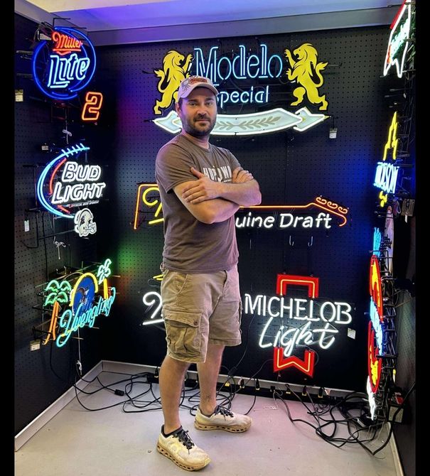 Man standing in a room with illuminated neon signs, including beer brands. He has his arms crossed.