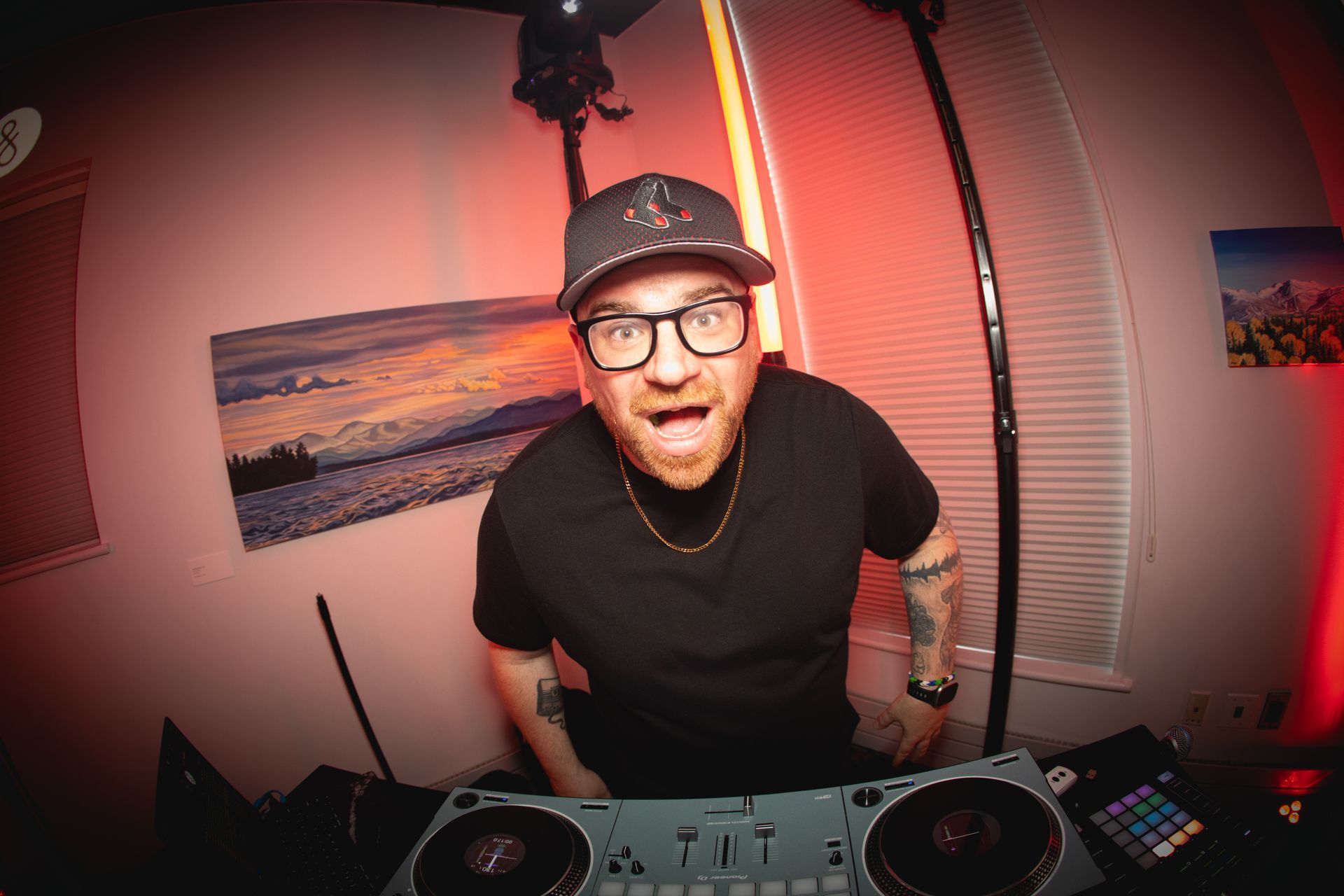Man in camouflage cap, glasses, and headphones points at the viewer, standing in front of a DJ booth in a brick-walled space.