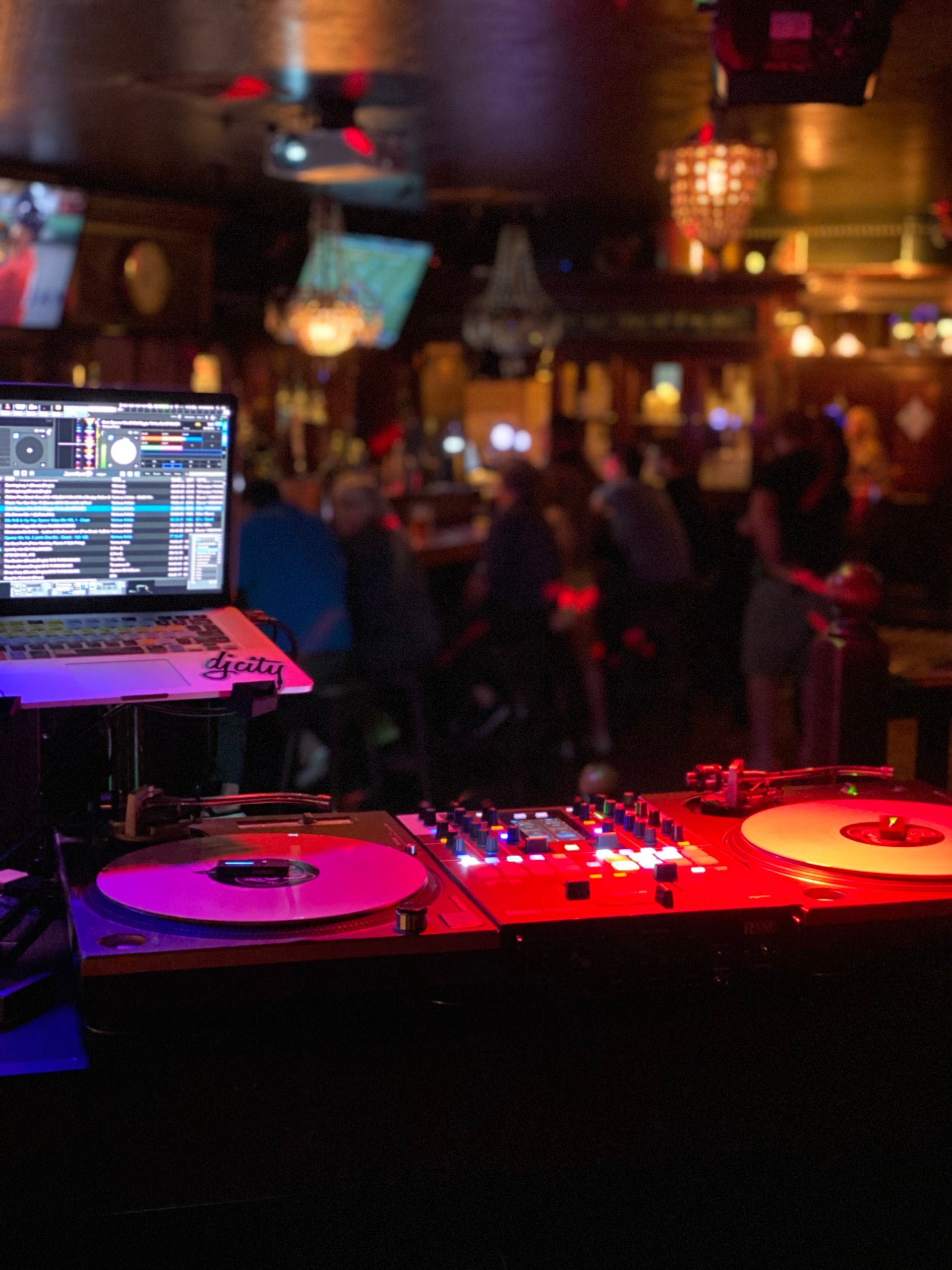 DJ setup in a dimly lit bar, with laptop, turntables, and mixer illuminated by purple and red lights.