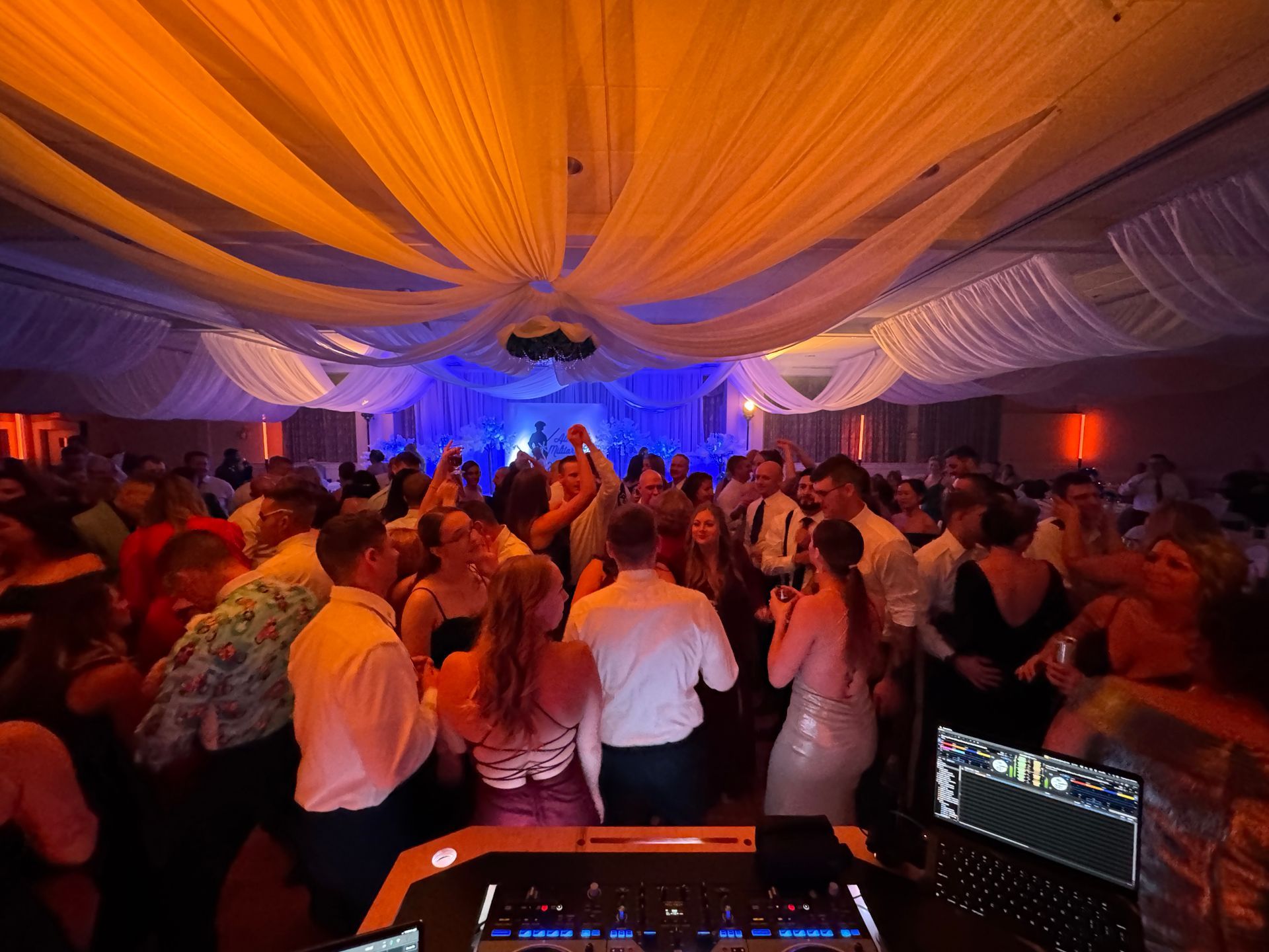 People dance at a party in a large room with fabric draped from the ceiling, lit with orange and blue lights.