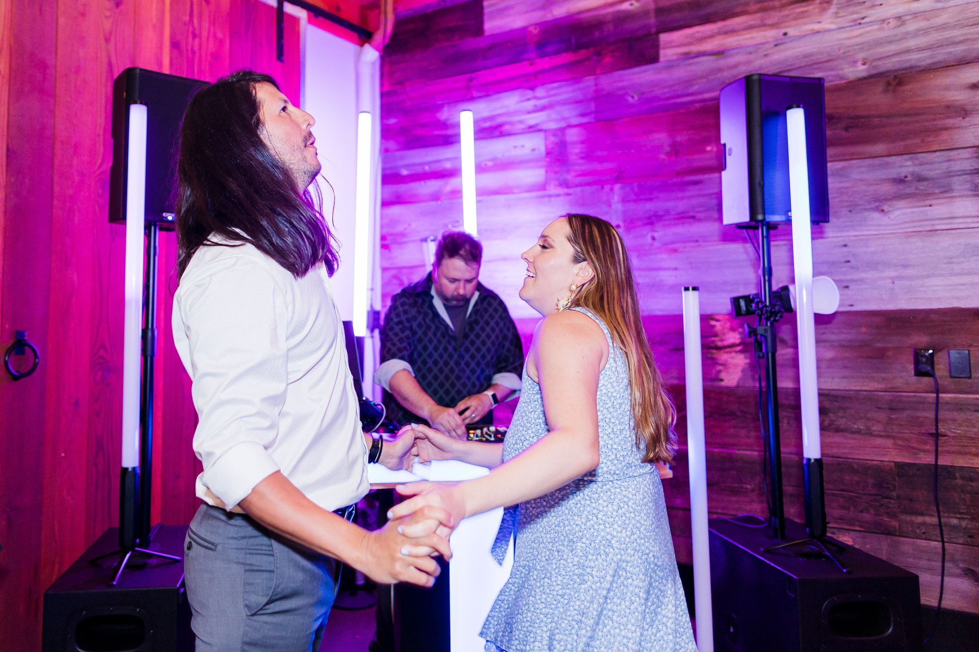 Two people dance at a party with a DJ.  Purple lighting, speakers, and wooden backdrop.