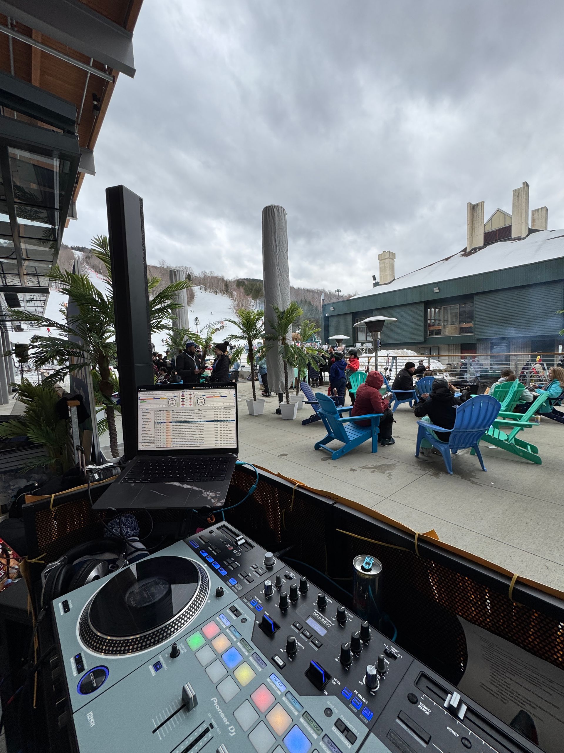 DJ setup outdoors with listeners in Adirondack chairs under a cloudy sky near a snowy landscape.