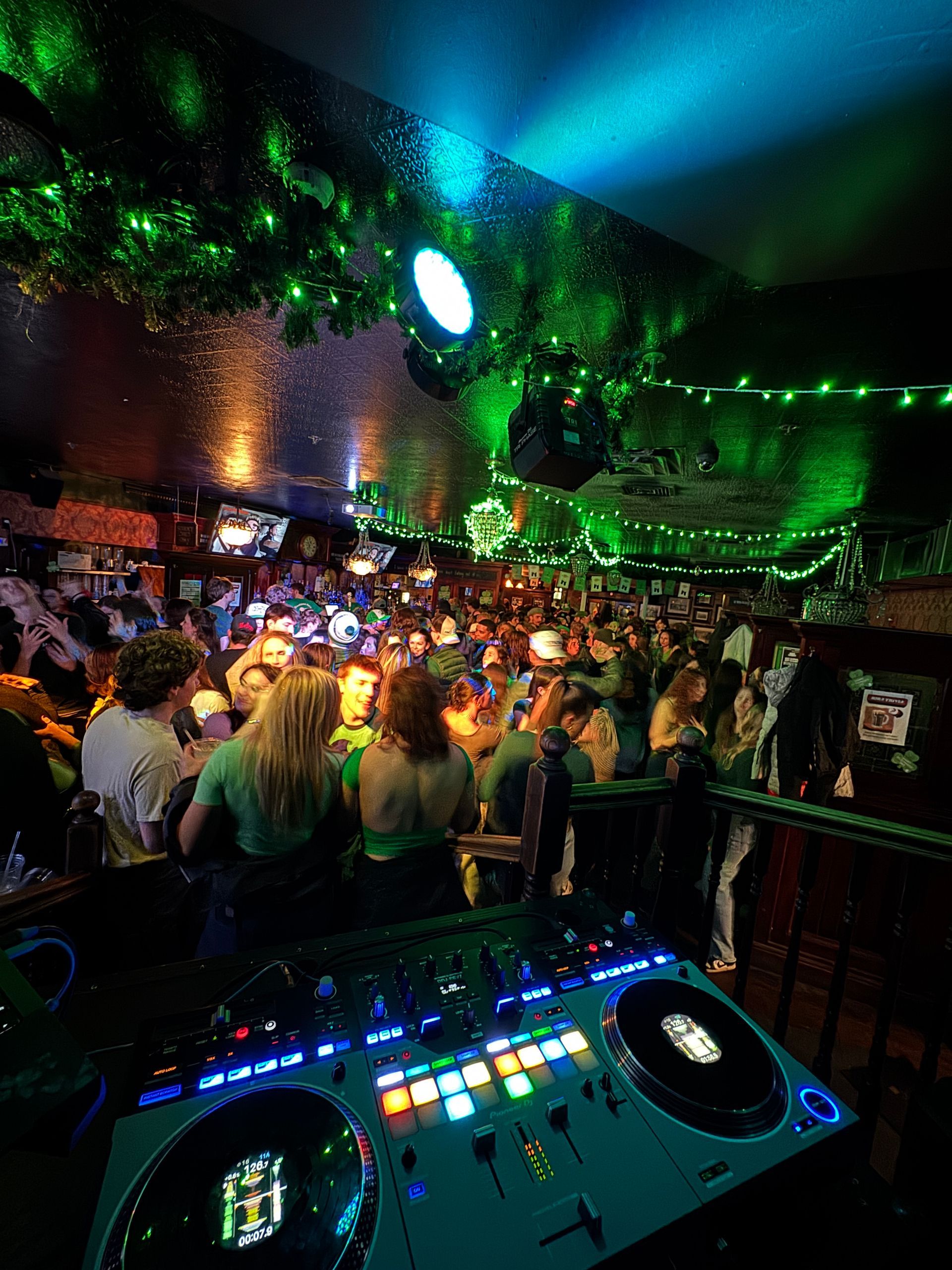 DJ setup with crowd in a dimly lit bar, green lights, people dancing.