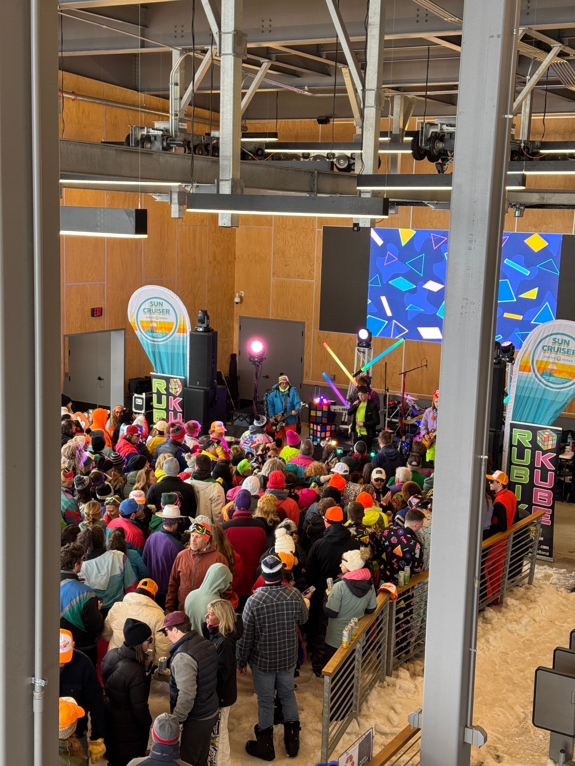 Large crowd at an indoor event with a band on stage, colorful clothing, and snow-like ground.