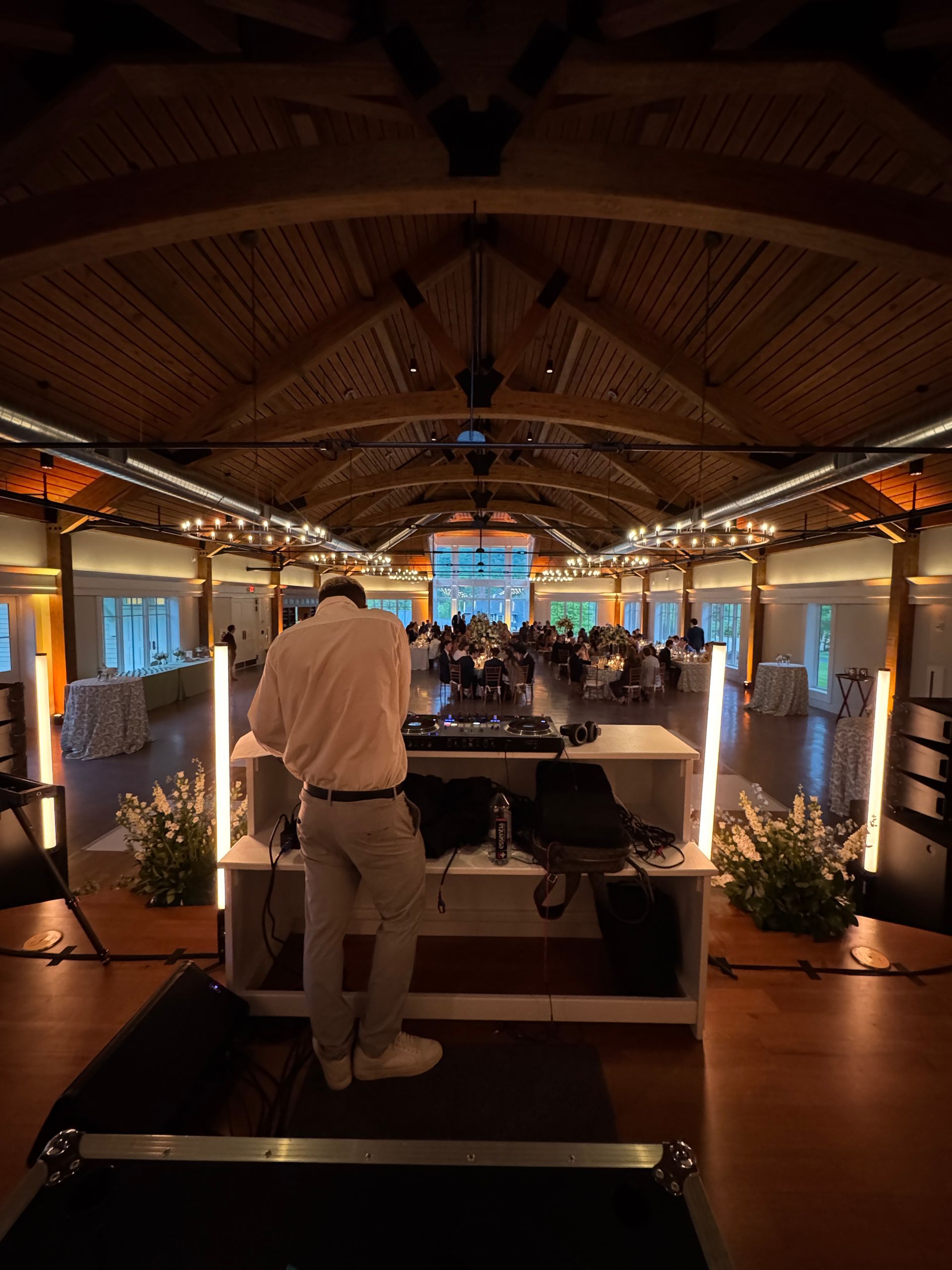 DJ at a wedding reception; indoor venue, wood ceilings, long tables, guests seated at tables, warm lighting.