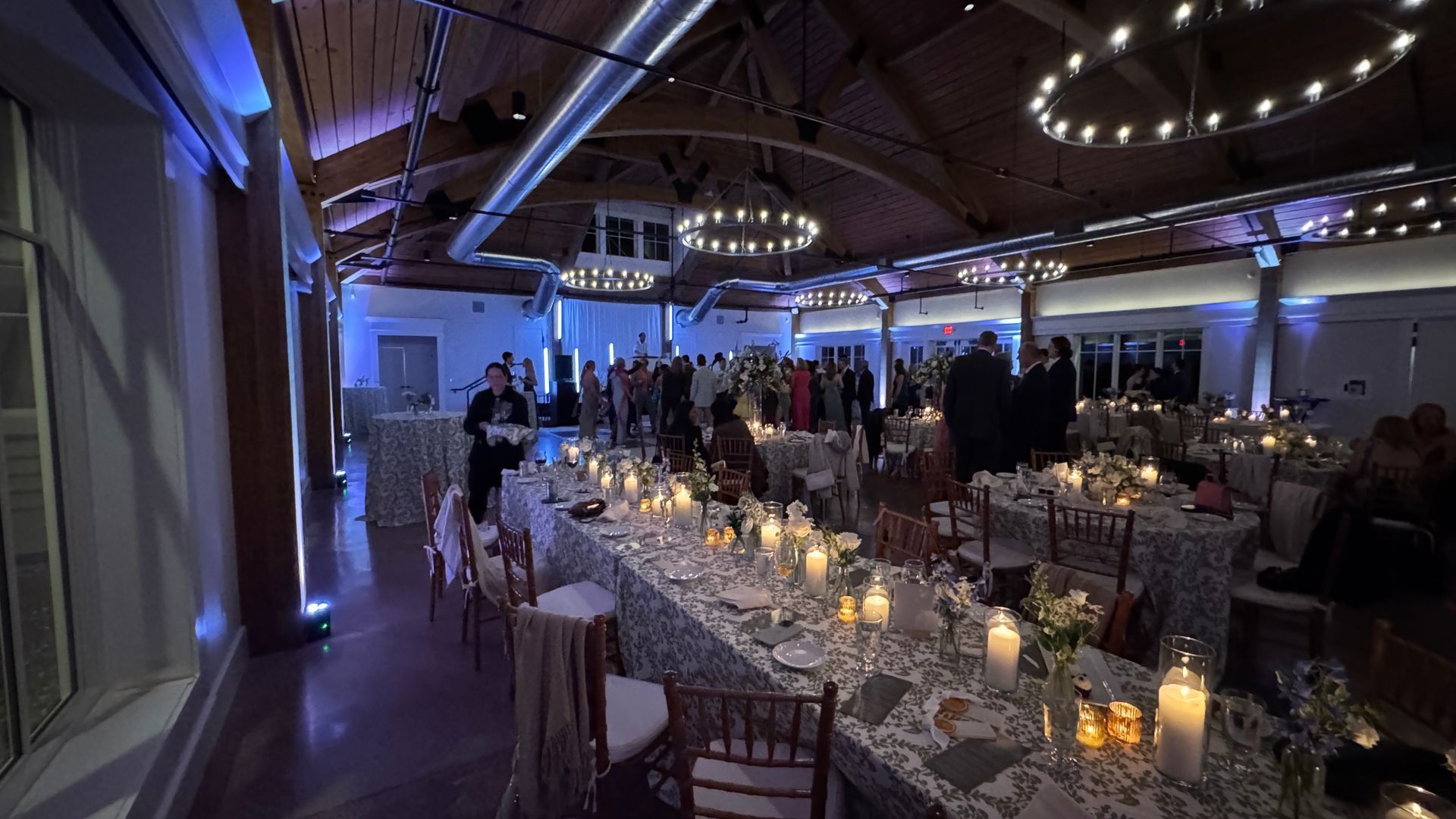 Reception hall with long tables set with candles, guests mingling, and overhead lighting.