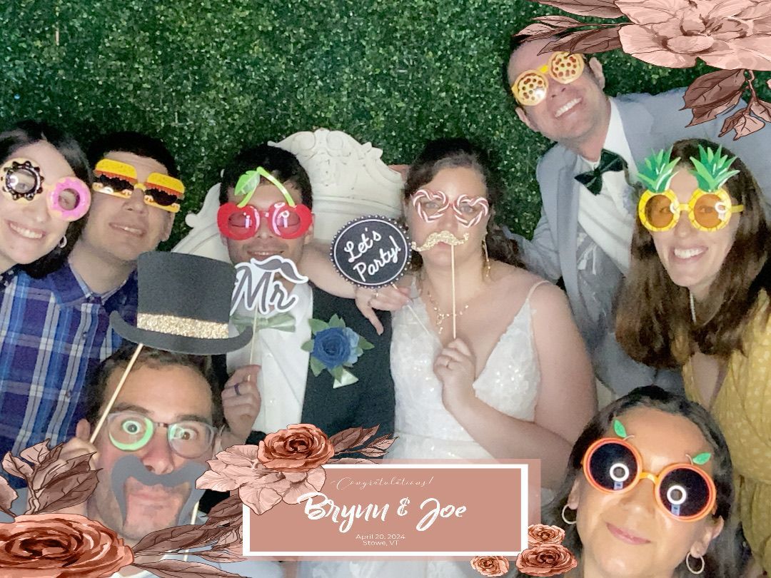 Group of people in photo booth with props, celebrating a wedding. Green background, flowers, and a banner.