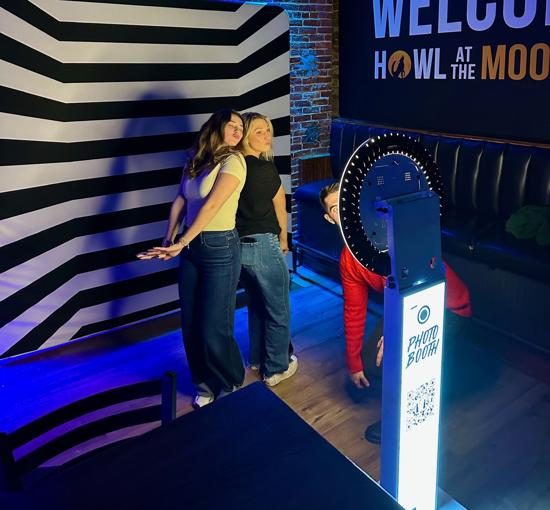 Two women pose for a photo with a photo booth at Howl at the Moon. One person kneels near the machine.