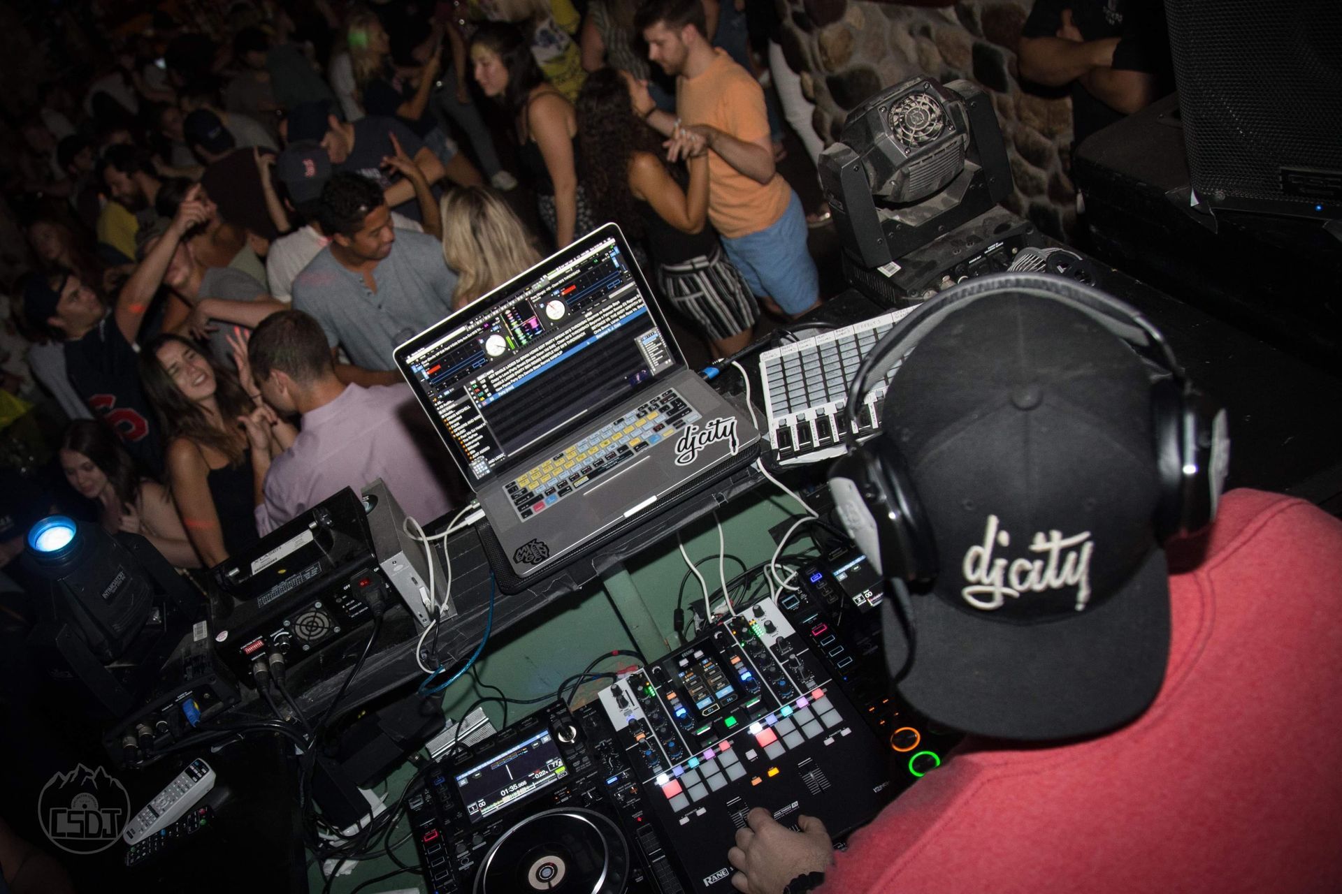 DJ in a dimly lit club setting, playing music with equipment, with a crowd of people dancing and enjoying the music.