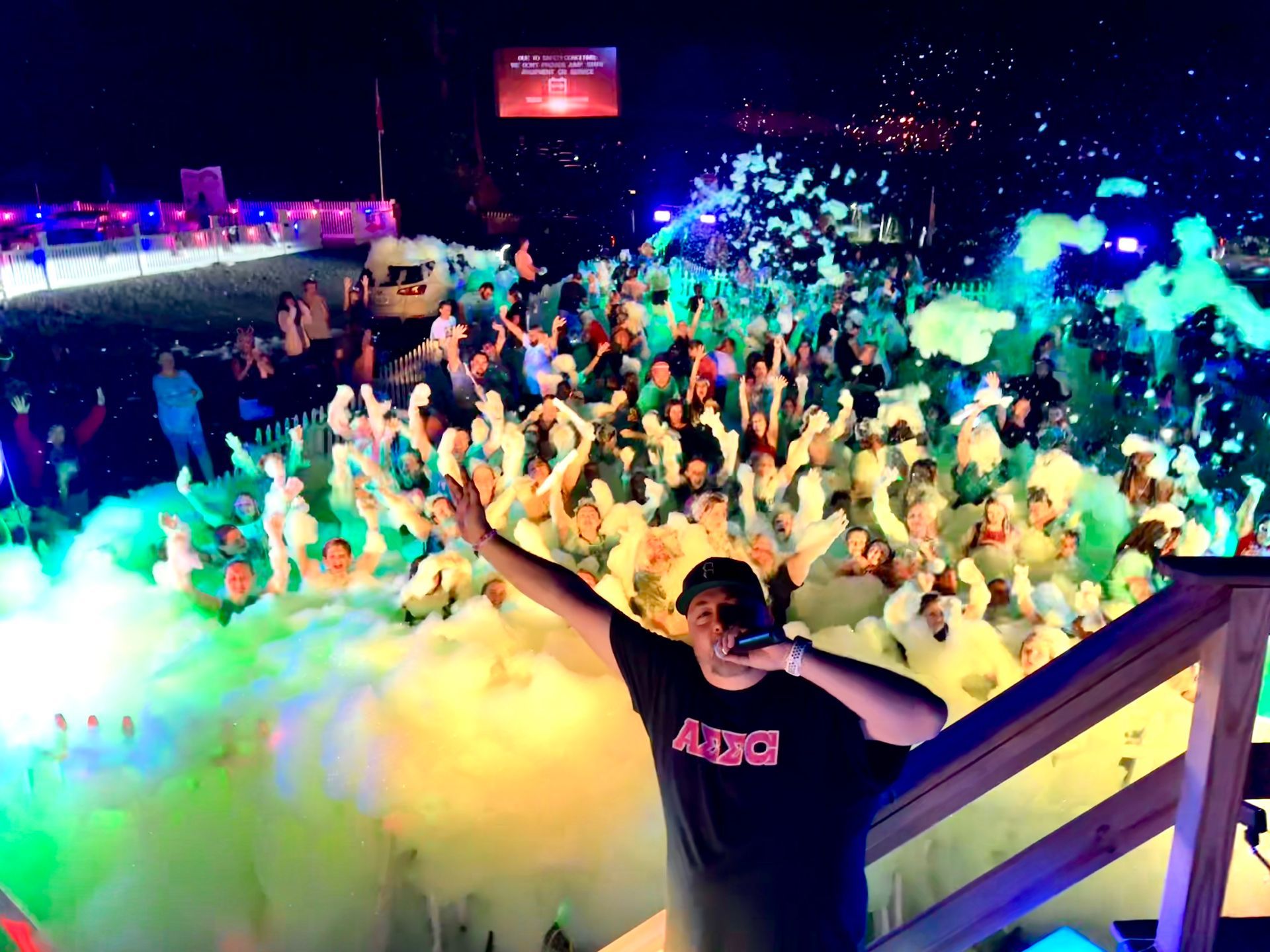 DJ performing at a foam party with crowd, lit with colorful lights.