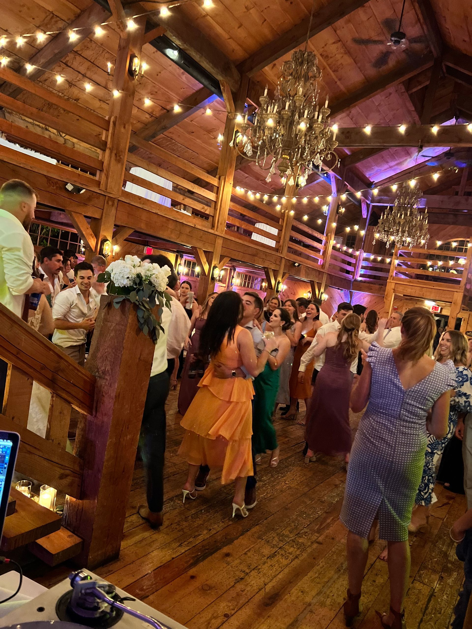 People dancing at a wedding reception in a wooden barn, with string lights and chandeliers.