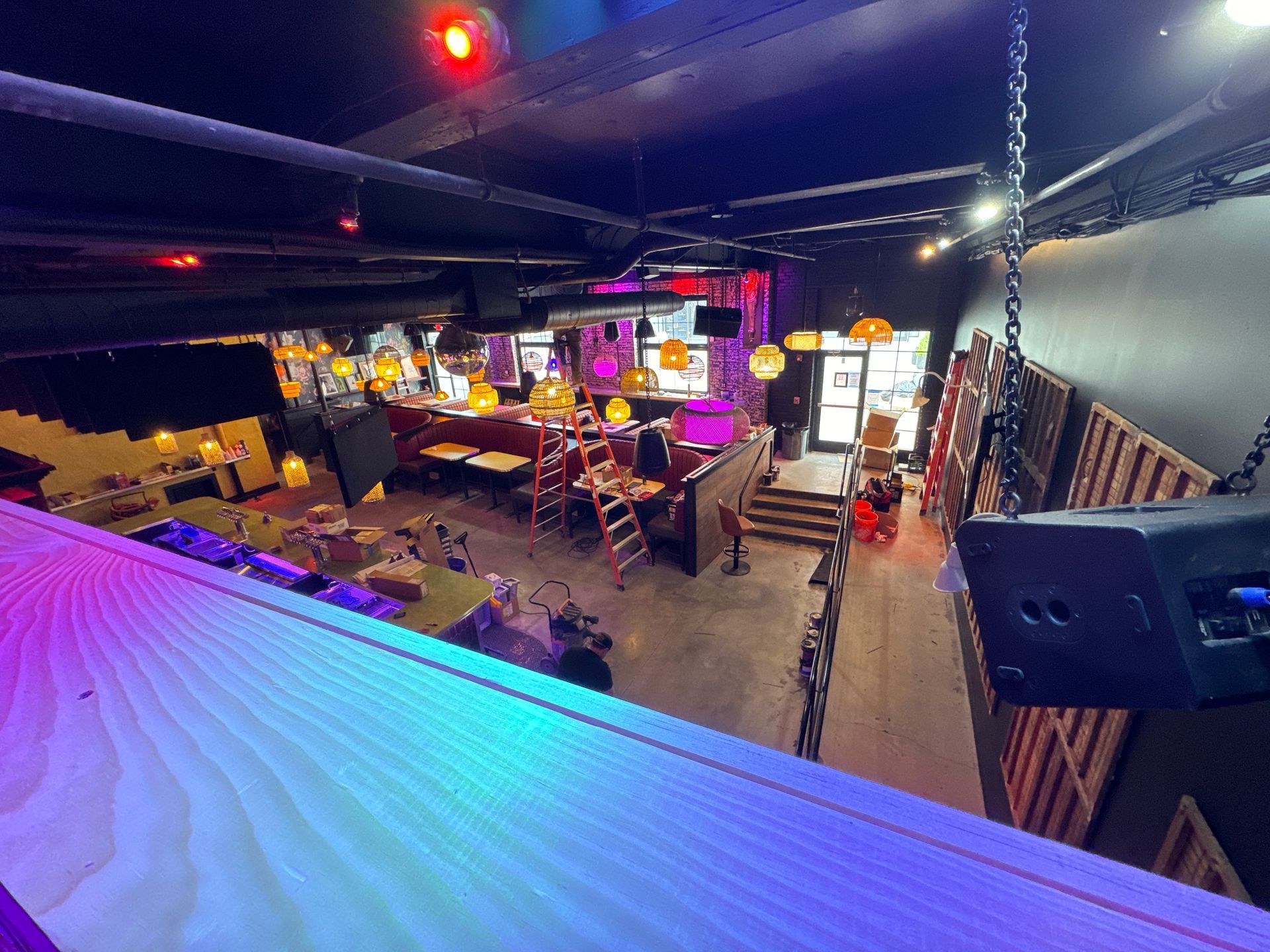 Interior of a dimly lit bar, view from above. Purple and turquoise lights illuminate seating, stage, and stairs.