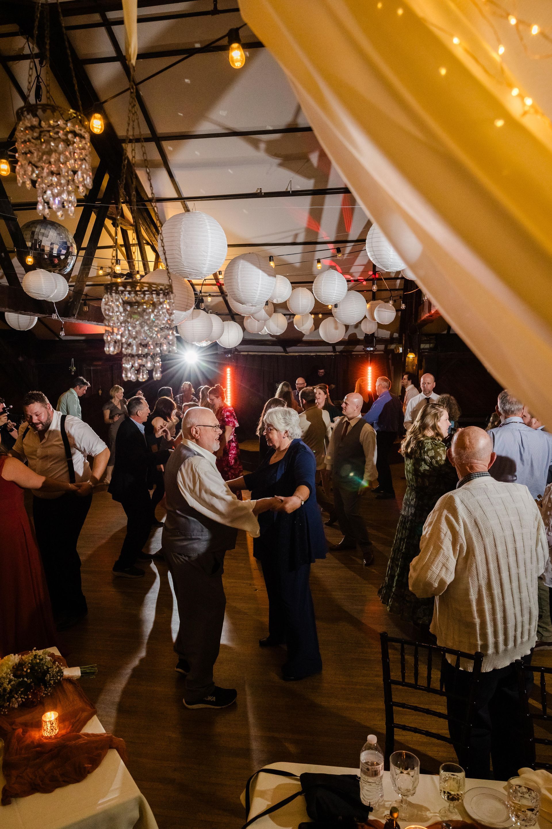 Wedding reception: Guests dance in a dimly lit industrial space with string lights, paper lanterns, and chandeliers.