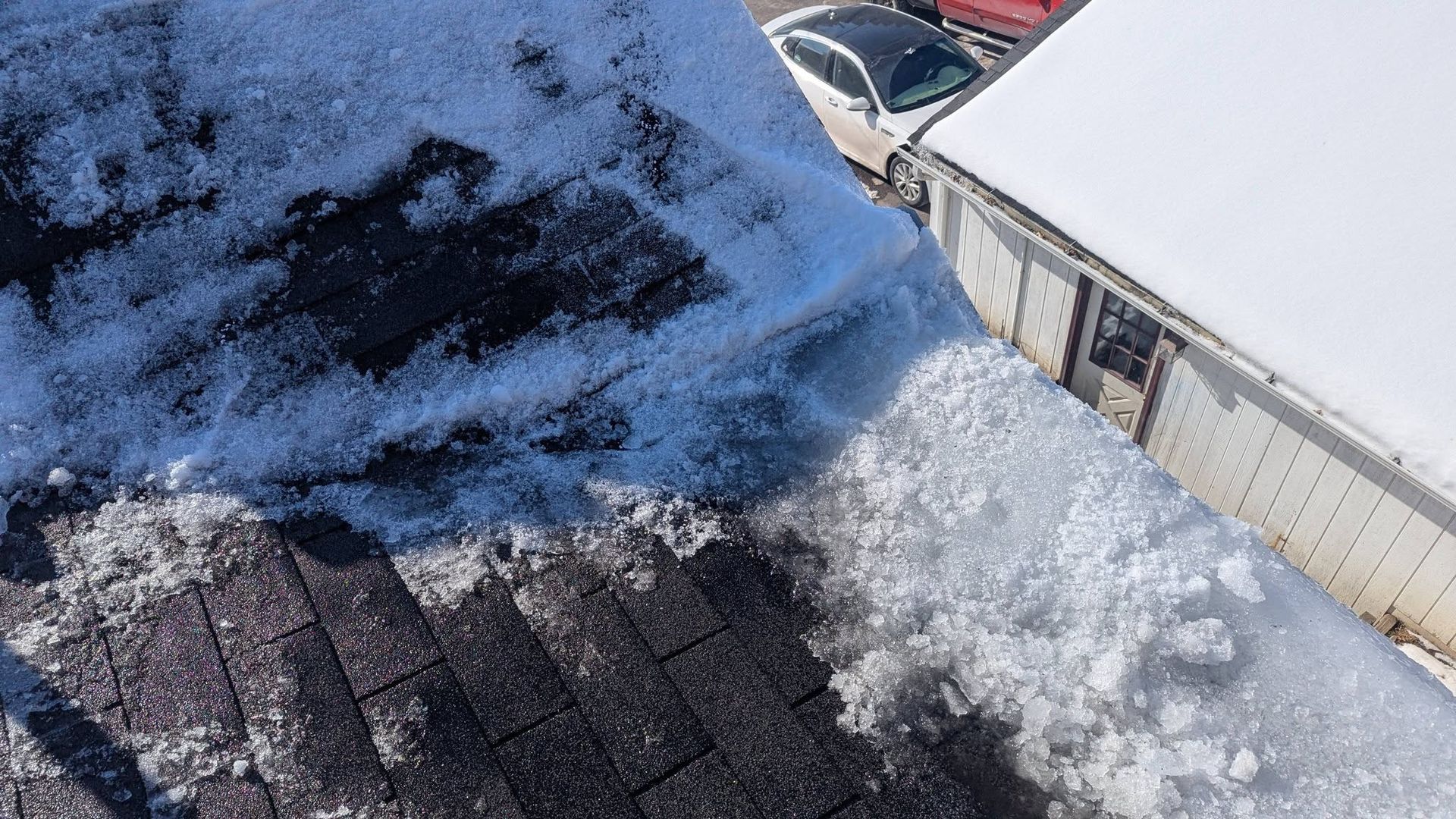 Snow-covered rooftop beside a white building, with a dark shingled roof and parked cars below.