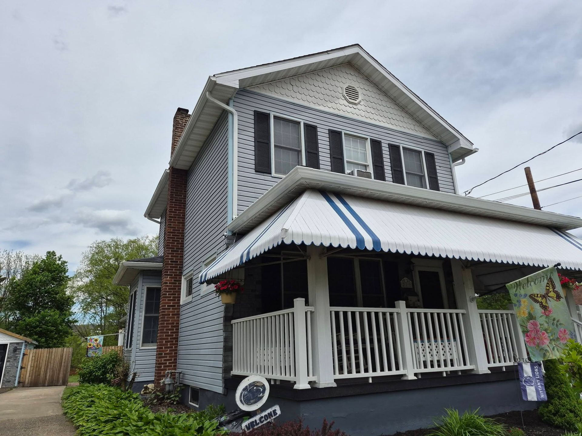 Two-story white house with porch, striped awning, and brick chimney on a cloudy day