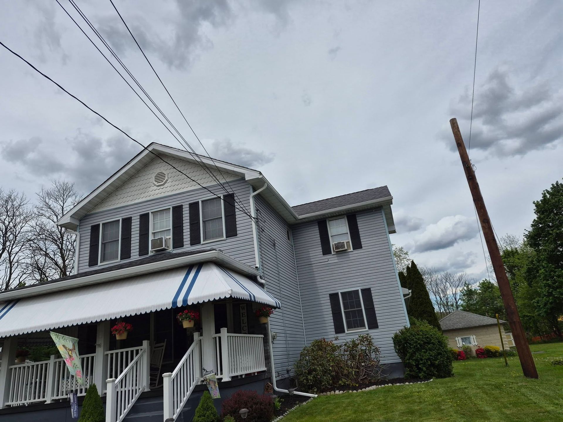 Gray two-story house with a striped porch awning under cloudy skies, viewed from the street.