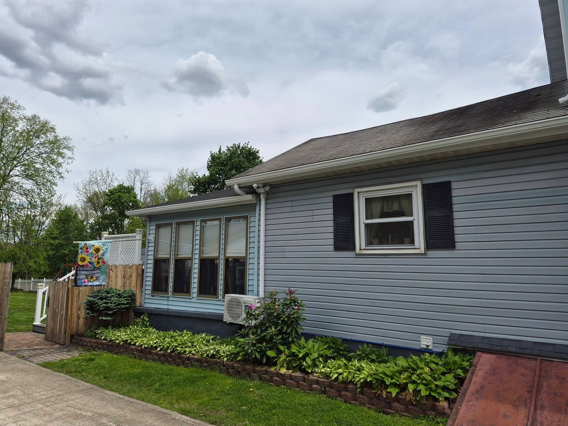 Single-story gray house with black shutters, small front garden, and cloudy sky.