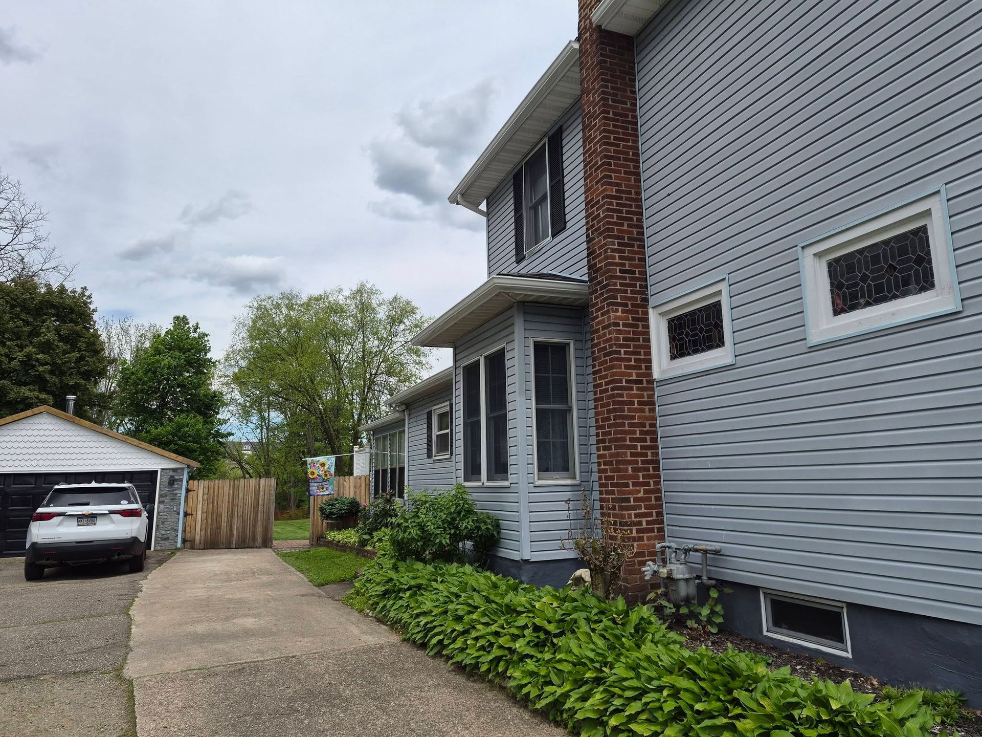 Side view of a gray house with brick chimney, driveway, garage, and green shrubs under cloudy sky