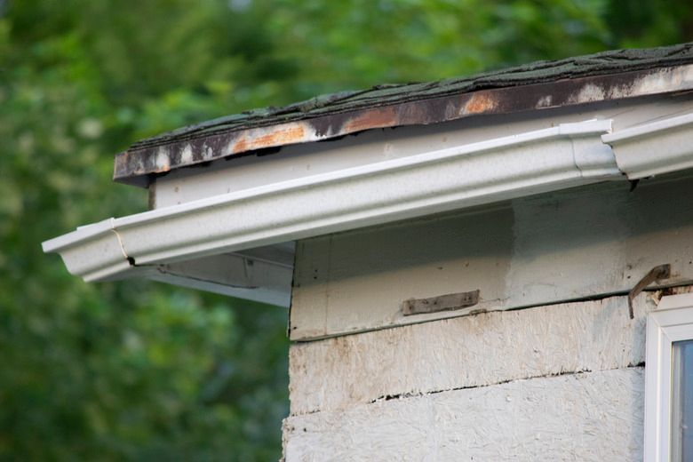 Damaged house roof edge with peeling trim and a loose gutter against a green background