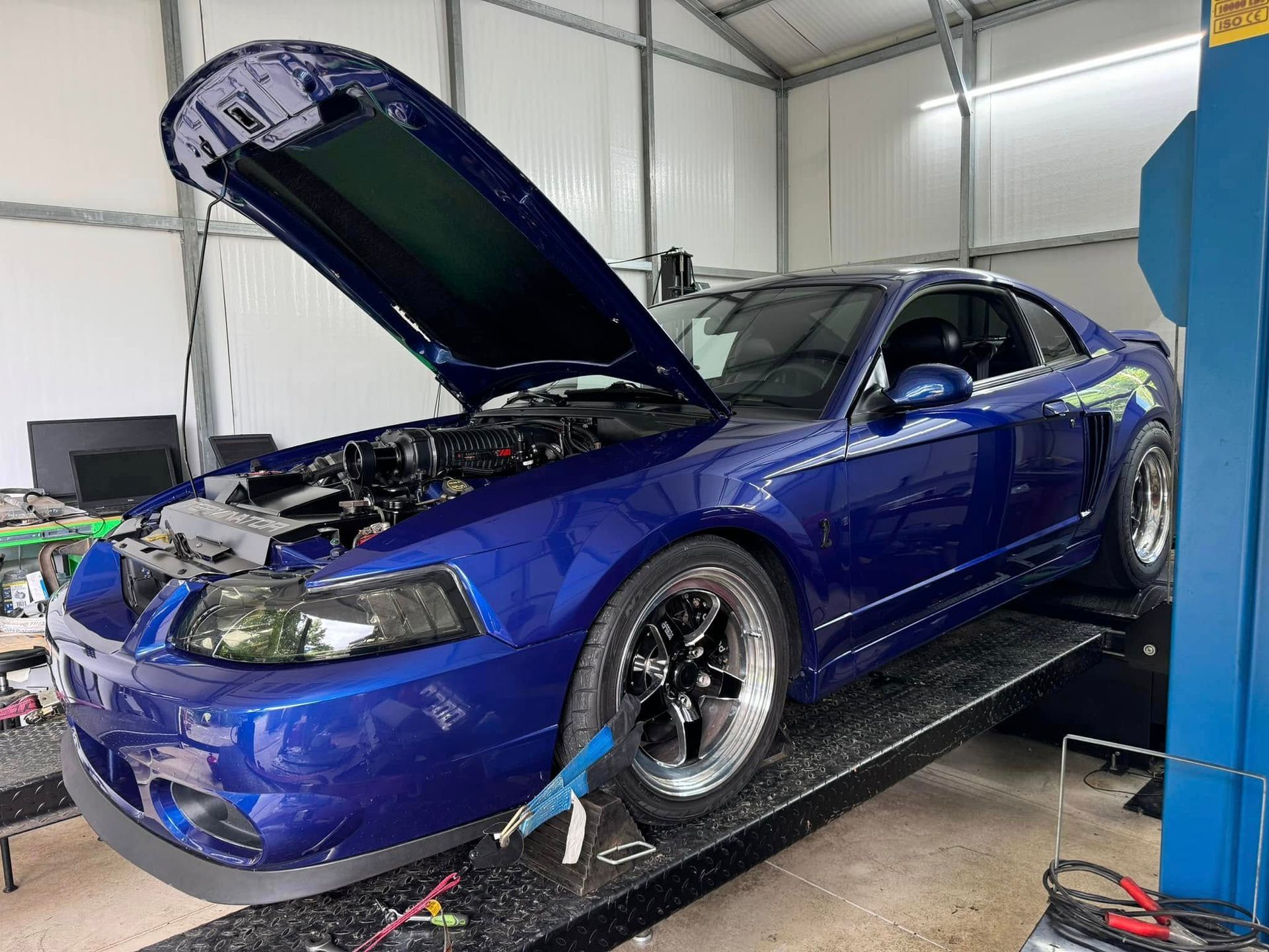 A blue mustang with its hood open is sitting on a lift in a garage.