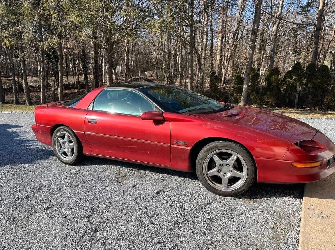 A red car is parked in a gravel lot in front of a forest.