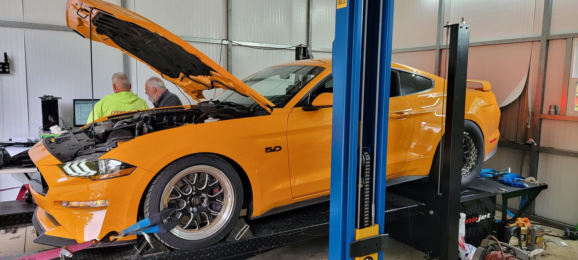 A yellow mustang is sitting on a lift in a garage with its hood open.