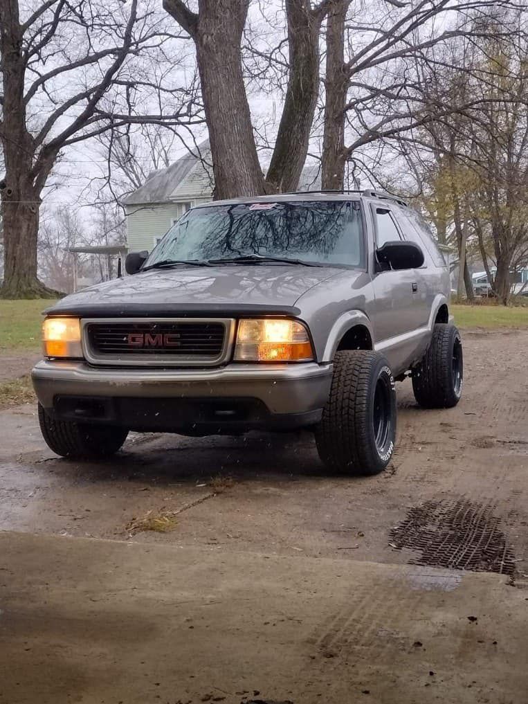 A gray gmc blazer is parked in a driveway next to trees.