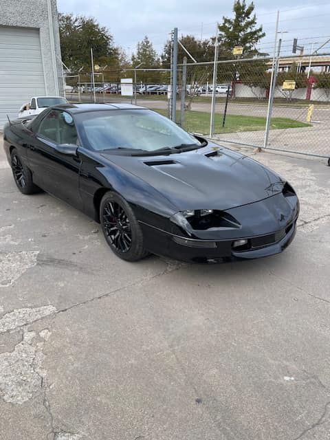 A black car is parked in a parking lot next to a fence.
