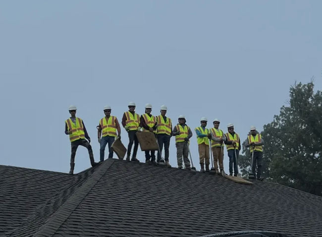 Construction workers on a roof, wearing hard hats and safety vests, against a cloudy sky.